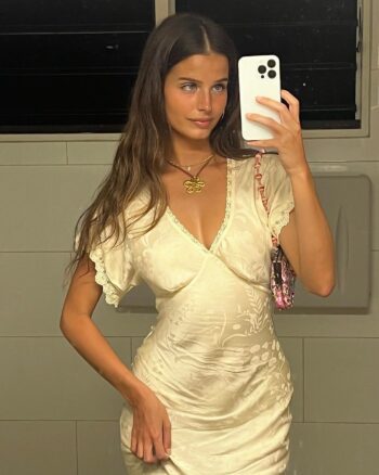 Hermosa modelo australiana Steph Mandich posando con un vestido blanco floral, collar dorado y cabello ondulado