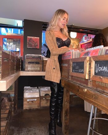 Influencer Corinna Kopf posando con lencería negra y abrigo beige en un entorno de discos y cajas de música