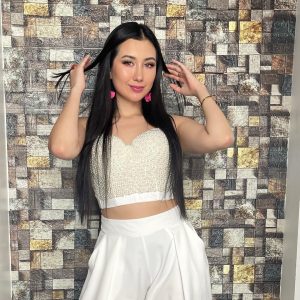 Kelly Bustamante posa con confianza en un fondo de pared de piedra, luciendo un crop top blanco brillante y una falda corta