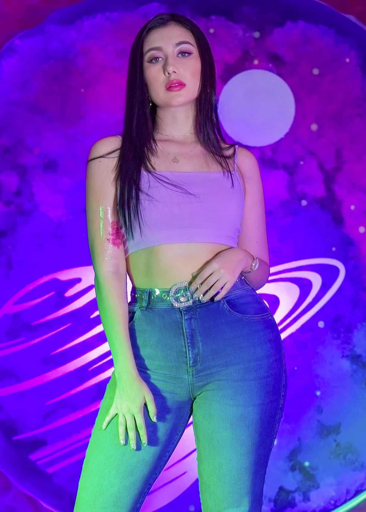 Kelly Bustamante posa con confianza en un ambiente púrpura, luciendo un top corto y jeans de tiro alto