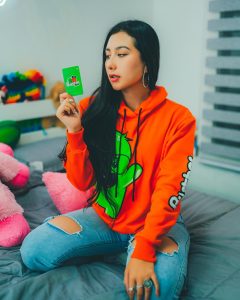 Kelly Bustamante posa con una sudadera naranja vibrante y jeans rasgados, mostrando su estilo fresco y desenfadado