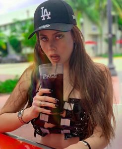 Ignacia Michelson sujeta una bebida de Coca-Cola con expresión seria, vistiendo minifalda negra y gorra de béisbol LA