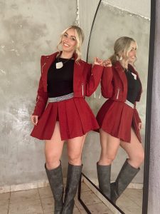 Natalia Rivera sonriendo con estilizado atuendo rojo y botas de cuero gris, posando junto a su compañera