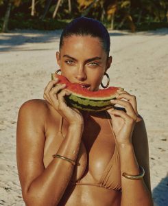Priscilla Huggins Ortiz con traje de baño rojo mordiendo una rebanada de sandía, posando sonriente en una playa tropical