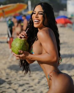Yarishna Ayala sonriendo mientras sostiene un coco verde en la playa
